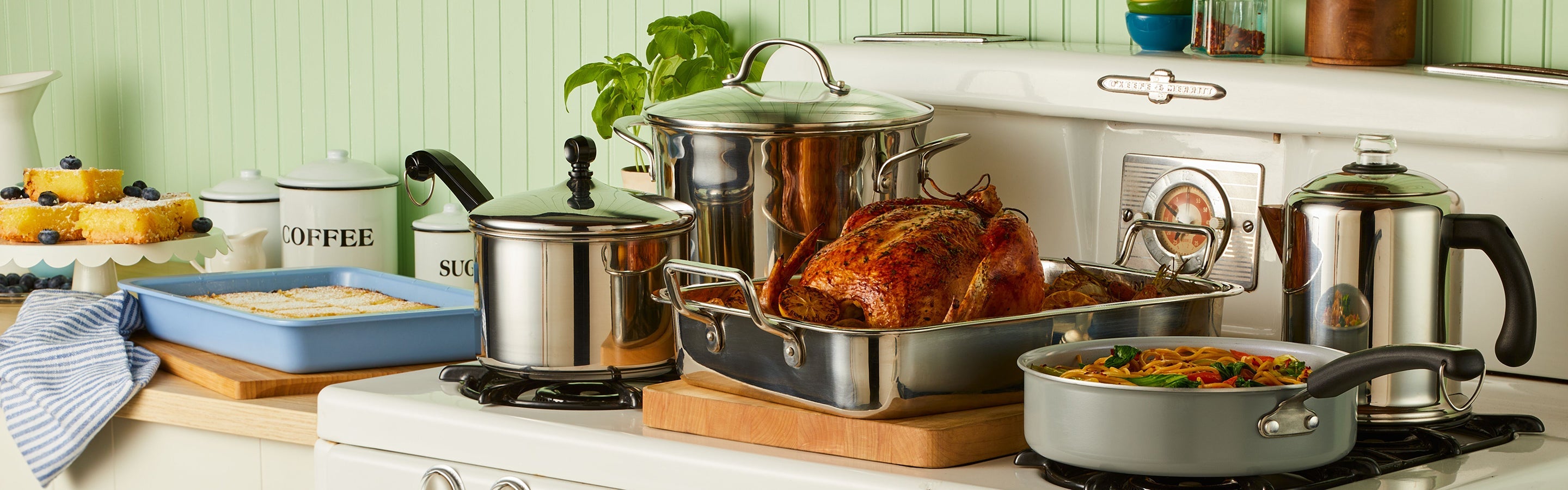 A kitchen scene with various stainless steel cookware on a stove, including a roasting pan with a cooked turkey, saucepans, a skillet with pasta, and a coffee percolator.