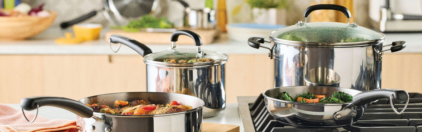Four stainless steel pots and pans with black handles, filled with various cooked foods, are arranged on a stovetop in a modern kitchen setting.