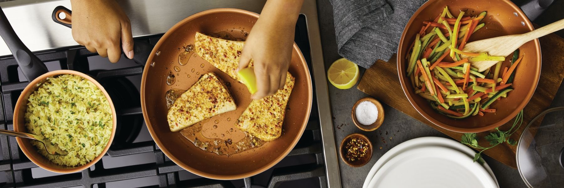 A person's hand squeezing lemon over fish fillets in a pan. Nearby, there is a pot of rice and a pan with sautéed vegetables on a stove.