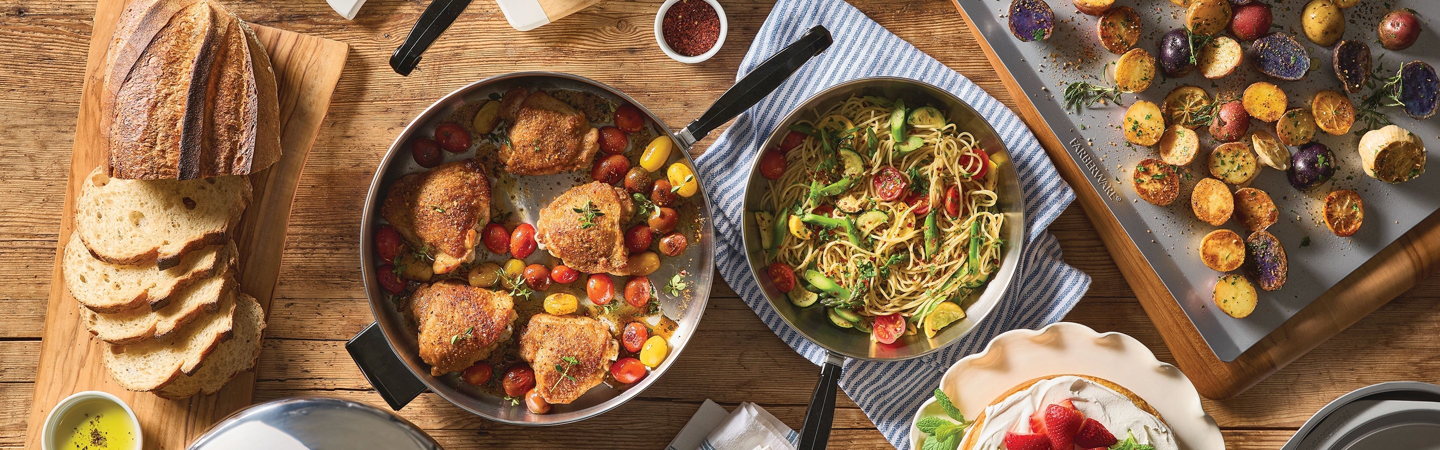 An overhead view of a wooden table set with sliced bread, sautéed chicken with tomatoes, spaghetti with vegetables, and roasted mixed potatoes.