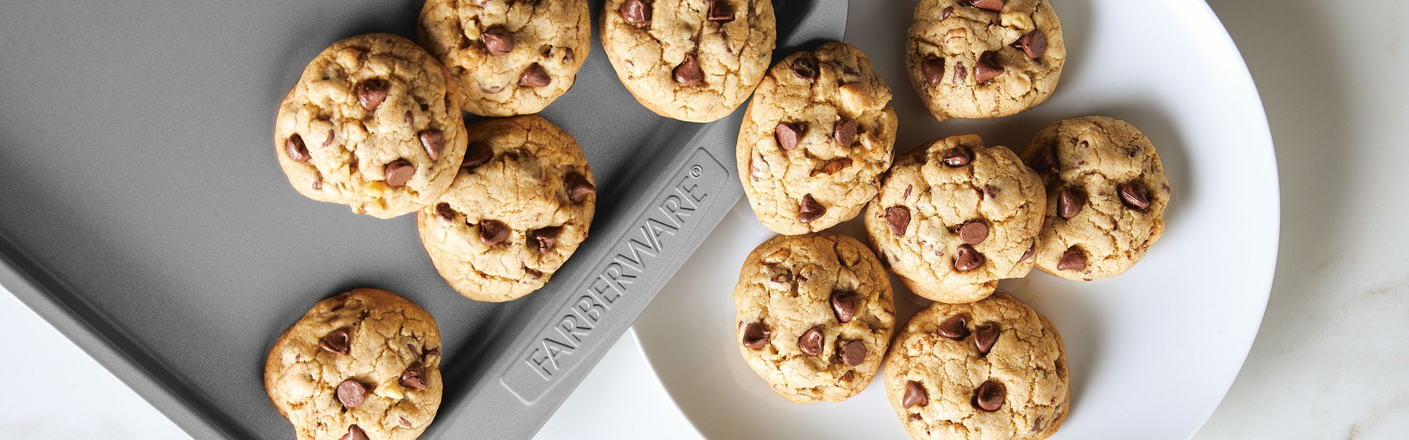 Chocolate chip cookies on a white plate and a gray Farberware baking sheet, viewed from above.