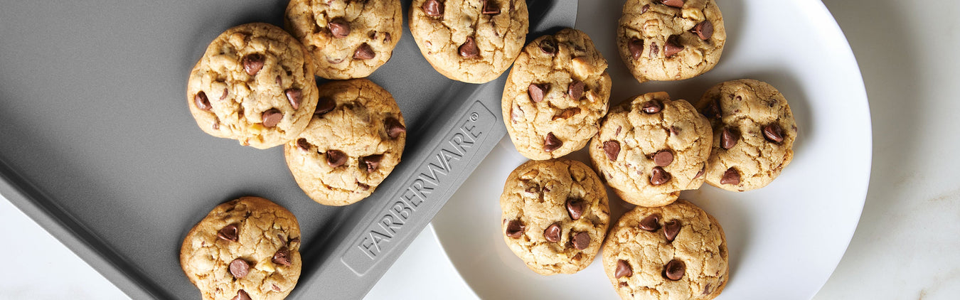 Chocolate chip cookies on a white plate and a gray Farberware baking sheet, viewed from above.
