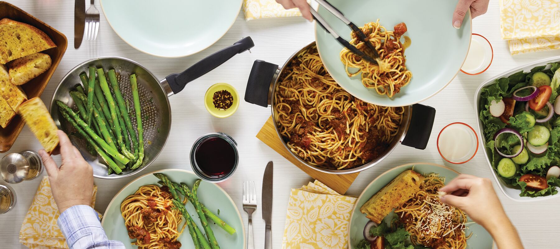 A table set with pasta, asparagus, salad, garlic bread, red wine, and a hand serving spaghetti with tongs.