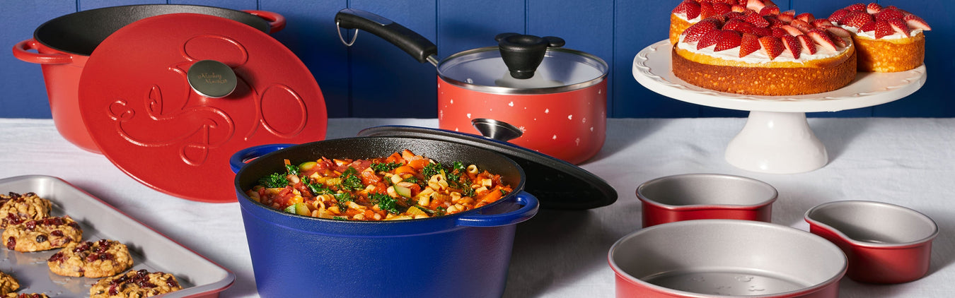 A variety of colorful cookware and bakeware with food, including a blue and red Dutch oven, saucepan, cookies, and cakes displayed on a white tablecloth.