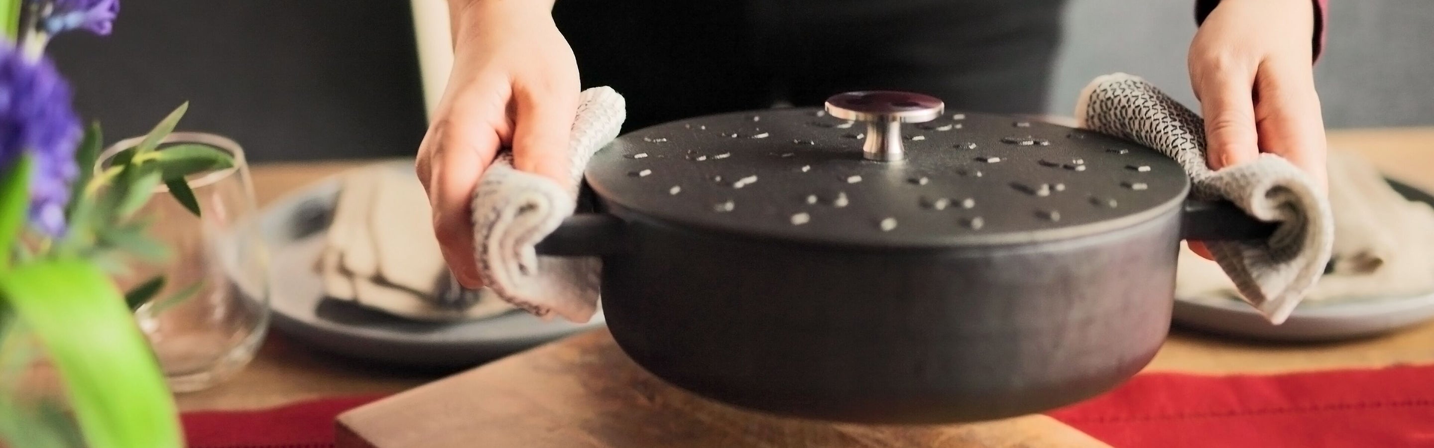A person holds a large black cast iron pot with a lid, using dish towels, over a wooden table set with plates and glasses.