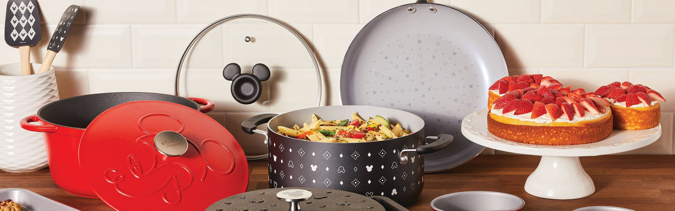 A kitchen counter displays Mickey Mouse-themed cookware, a pasta dish, a strawberry-topped cake on a stand, and various cooking utensils against a white tile backsplash.