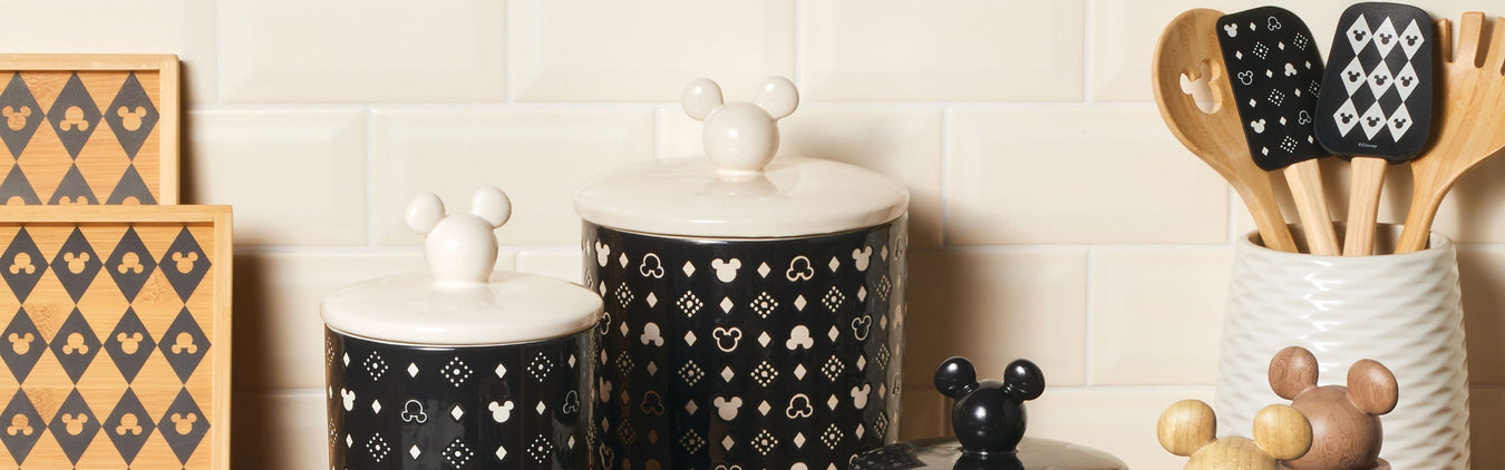 Black and white kitchen canisters, utensils, and cutting boards featuring Mickey Mouse patterns are arranged on a counter against a cream tile backsplash.
