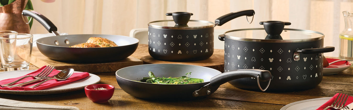 Three nonstick pots and pans with patterned exteriors sit on a wooden table, some containing food, next to plates with red napkins and cutlery.