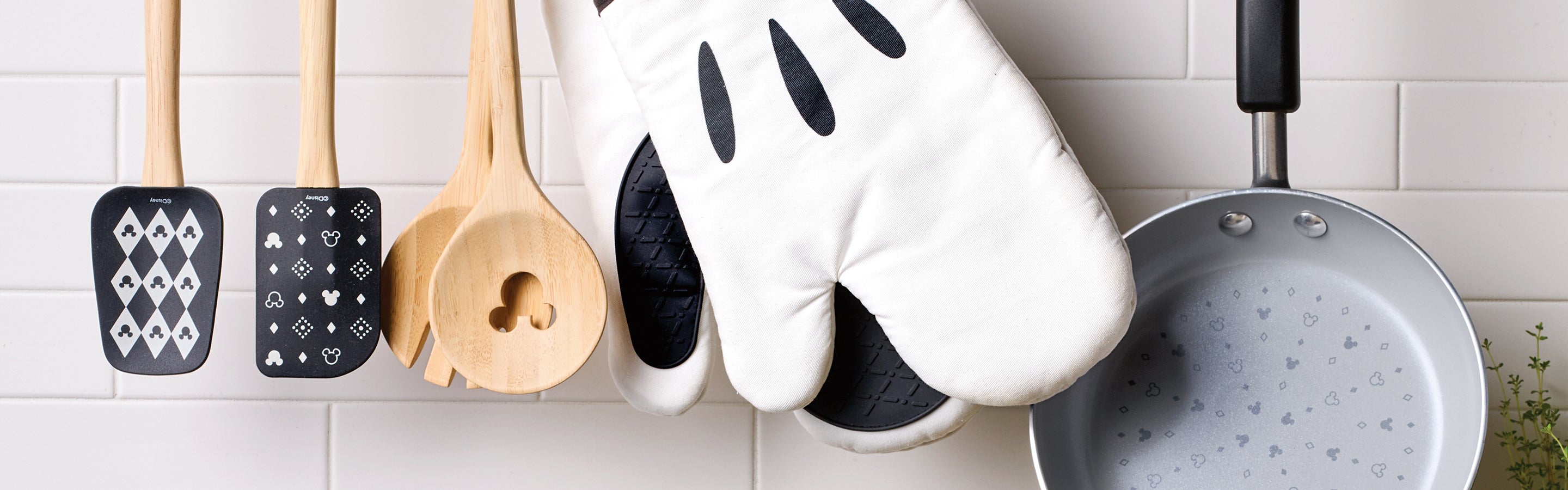 Kitchen utensils, including spatulas, wooden spoons, oven mitts, and a frying pan with card suit patterns, hang on hooks against a white tiled wall.