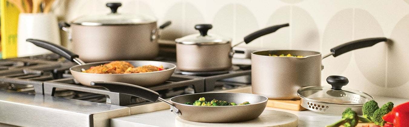 A set of pots and pans with black handles on a stovetop, some containing food, with vegetables and kitchen utensils nearby.