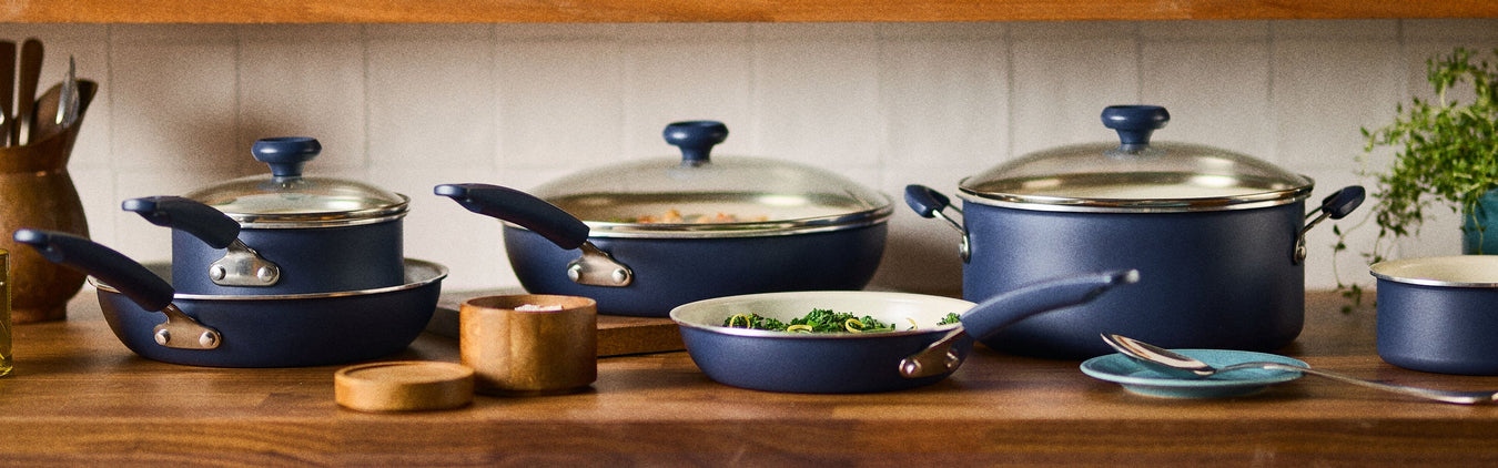A set of blue pots and pans with glass lids arranged on a wooden kitchen counter, with utensils, seasonings, and fresh greens visible nearby.
