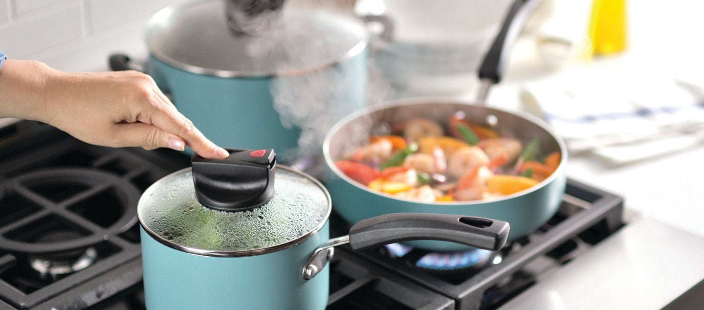 A hand holds the lid of a steaming teal saucepan on a stove, with a pan of mixed vegetables and seafood cooking on the adjacent burner.