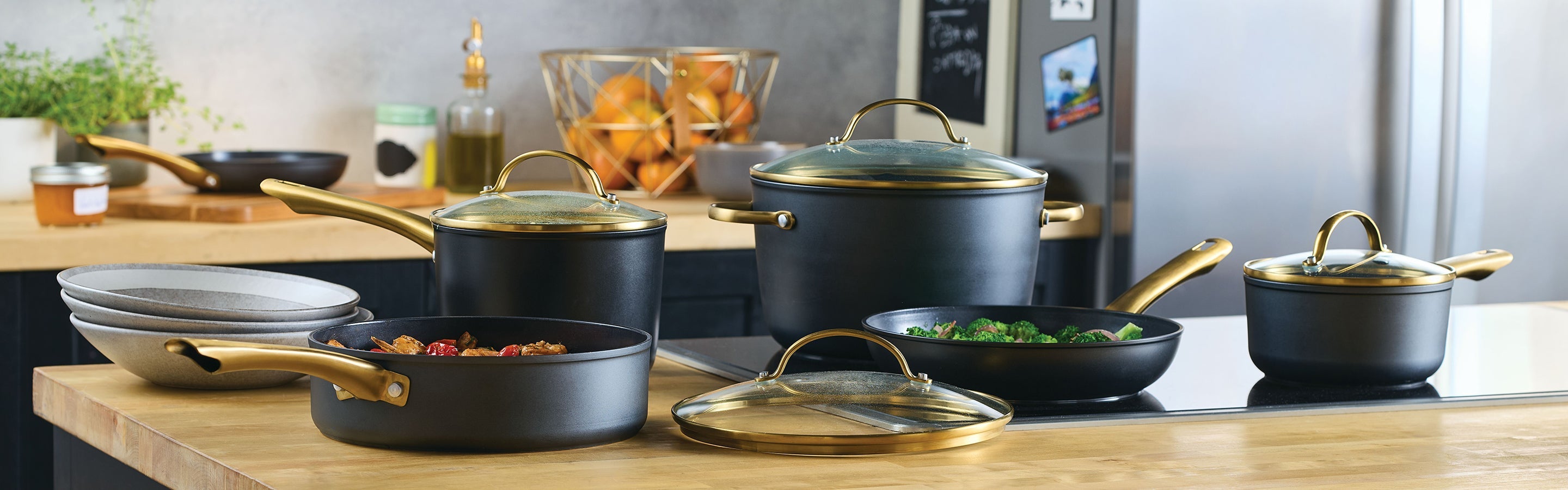 A set of black and gold pots and pans with glass lids is arranged on a kitchen counter, with bowls, vegetables, and utensils in the background.