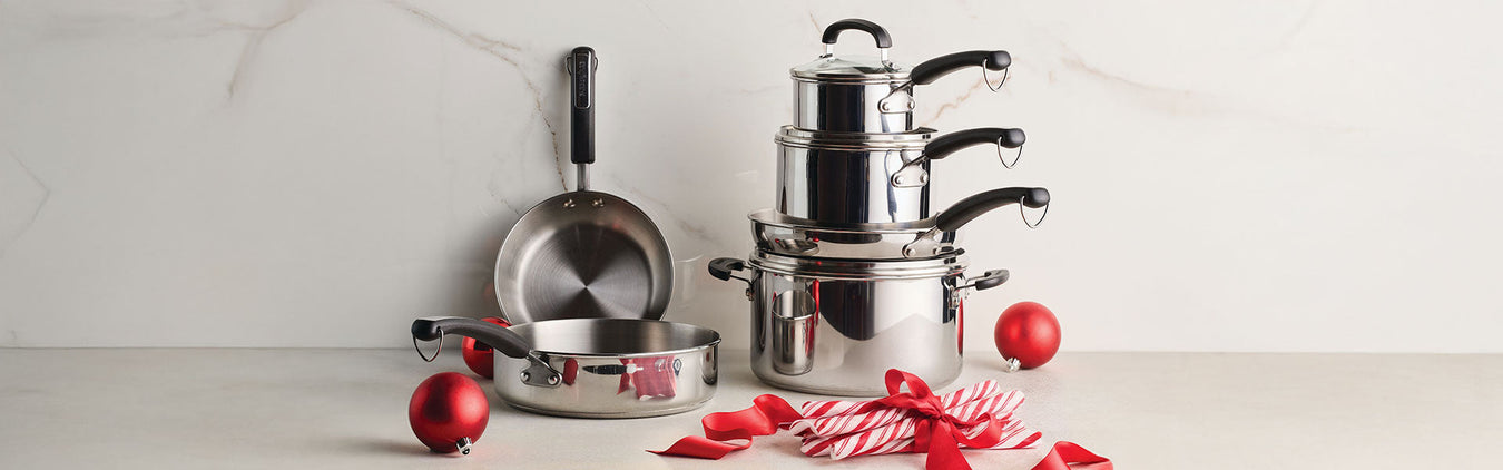 A set of stainless steel pots and pans stacked on a countertop, with red holiday ornaments and a ribbon in front.