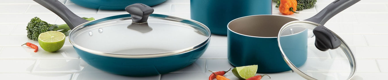 A set of teal nonstick cookware, including pans with glass lids, is displayed on a white tile surface with assorted vegetables and lime wedges.