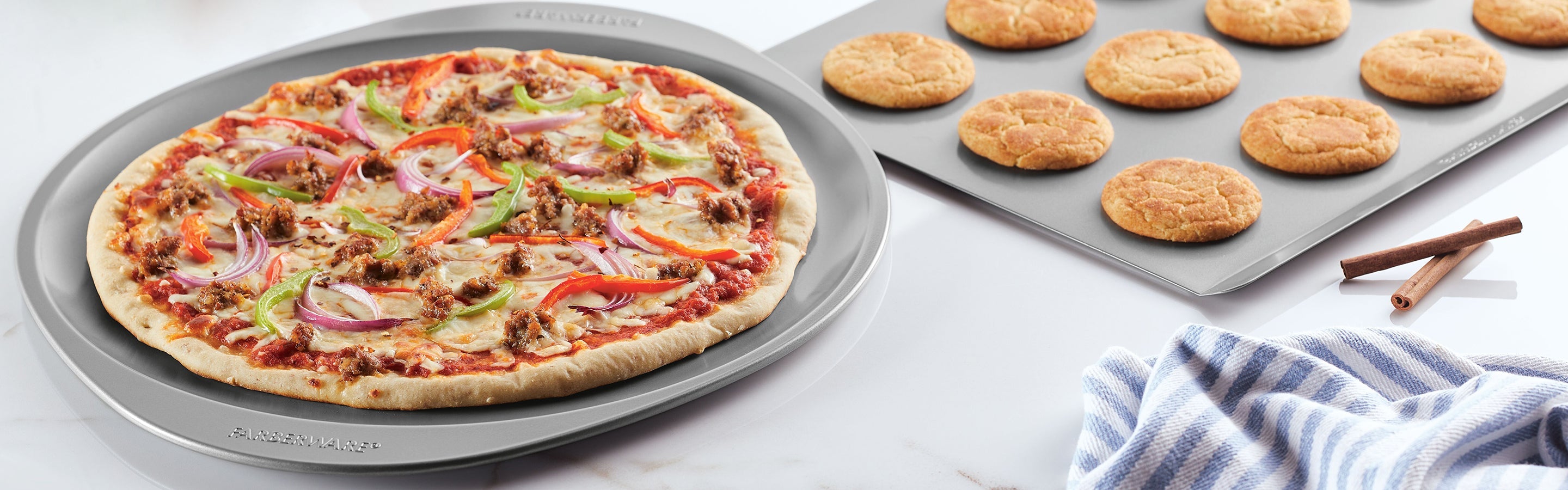 A round pizza with vegetables and sausage on a baking tray sits next to a tray of freshly baked cookies, with a striped towel and two cinnamon sticks nearby.