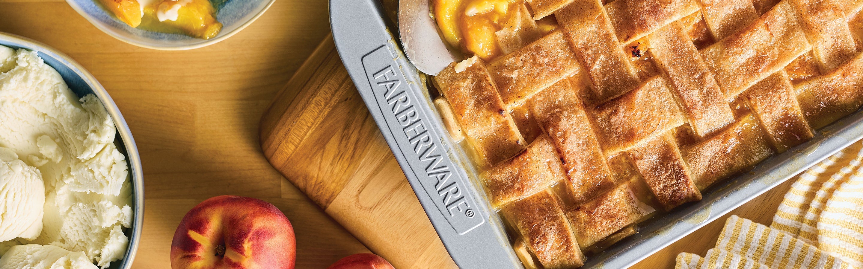 A baked peach pie with a lattice crust in a Farberware pan sits on a wooden table next to fresh peaches, a bowl of vanilla ice cream, and a yellow-striped towel.