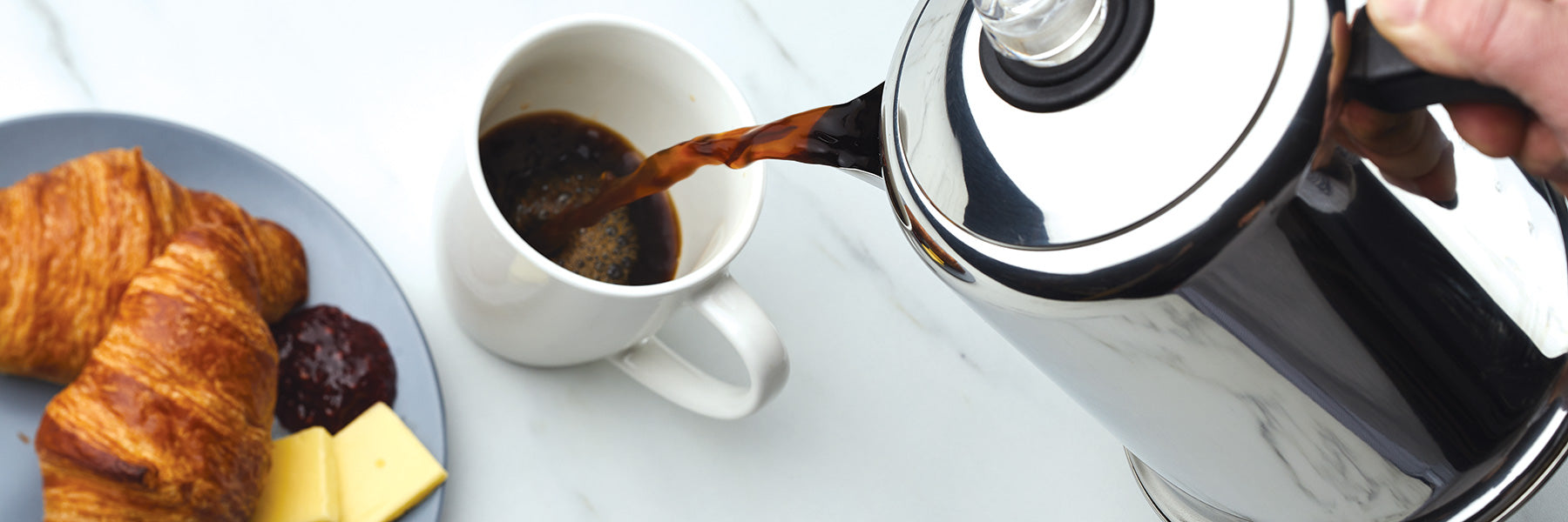 A person pours coffee from a French press into a white mug. Nearby are croissants, cheese, and jam on a plate.