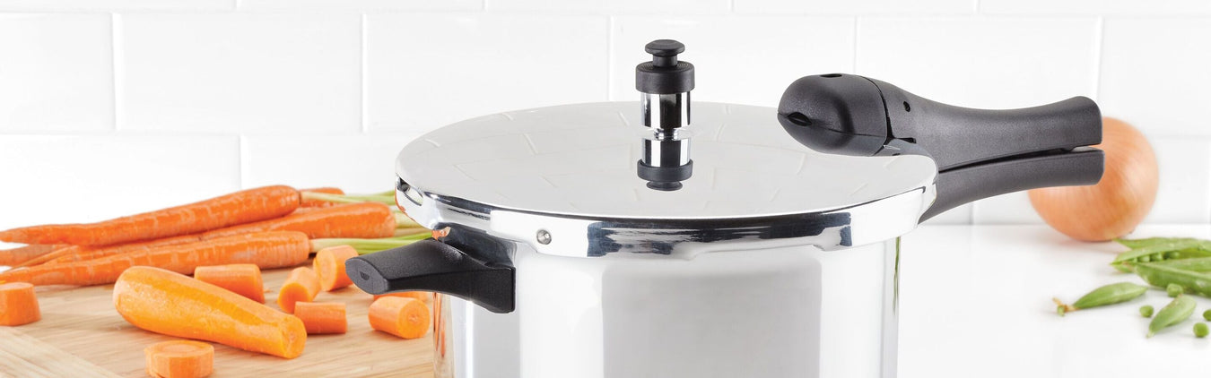 A stainless steel pressure cooker with black handles is on a counter near chopped carrots, whole carrots, green peas, and an onion, with a white tiled backsplash.
