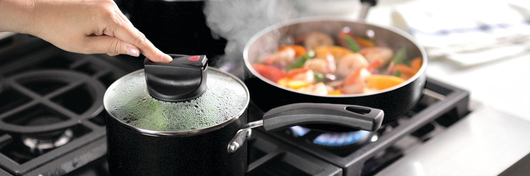 A person lifts the lid of a pot on a stove with steam rising. A pan in the background contains assorted vegetables and seafood, cooking over a flame.