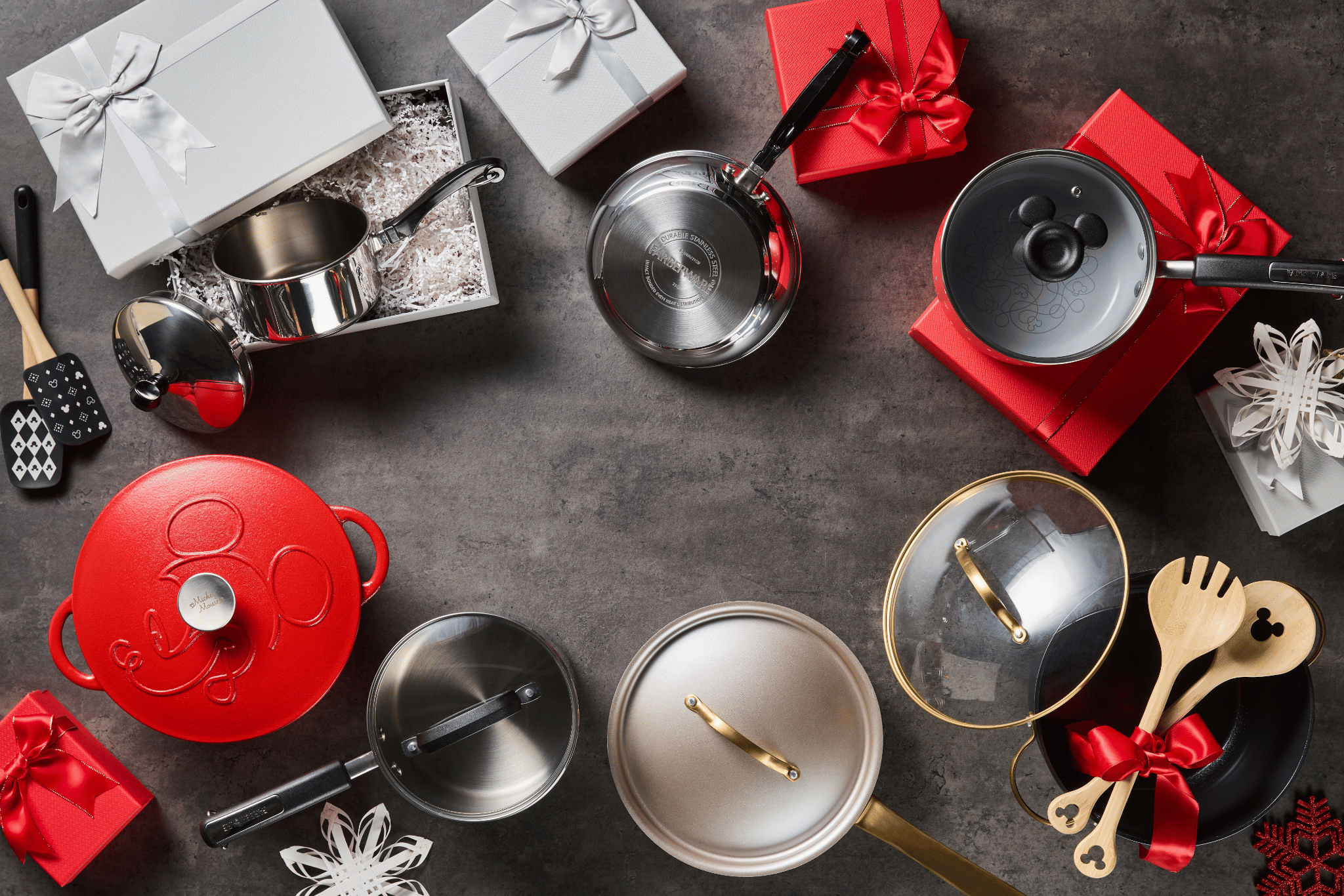 Assorted cookware, utensils, and gift boxes are arranged on a gray surface, including red and silver pots, pans, and wooden spoons with ribbon.