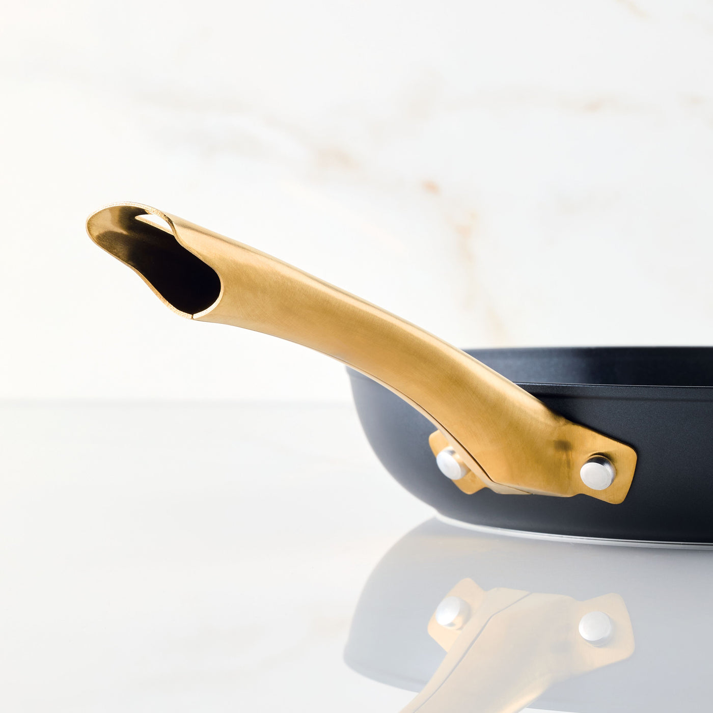 A close-up showcases the Forged Induction Ceramic 10-Inch Frying Pan with its gold-colored handle resting on a reflective surface. This nonstick marvel, made with forged aluminum and steel bases, guarantees even heat distribution and durability for all your culinary adventures.