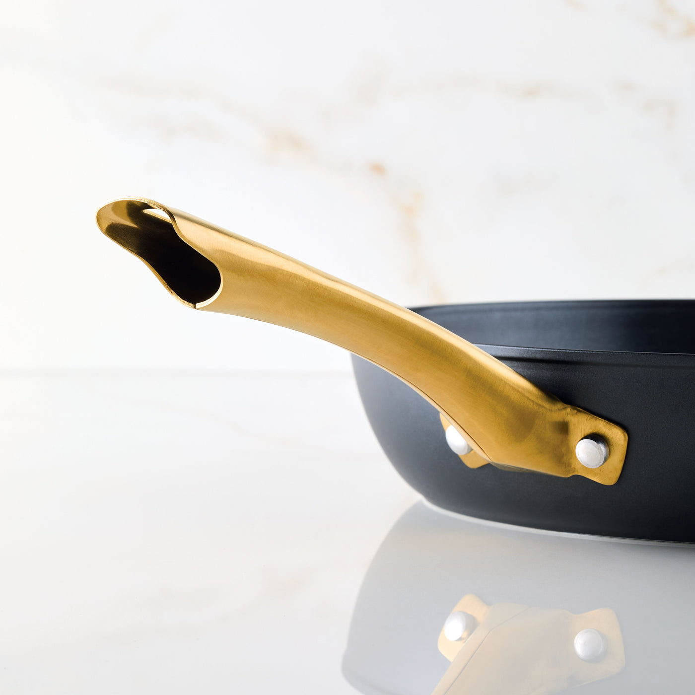 The Forged Induction Ceramic 12.25-Inch Frying Pan, featuring a gold handle and a black nonstick surface suitable for induction cooking, rests on a reflective marble surface.