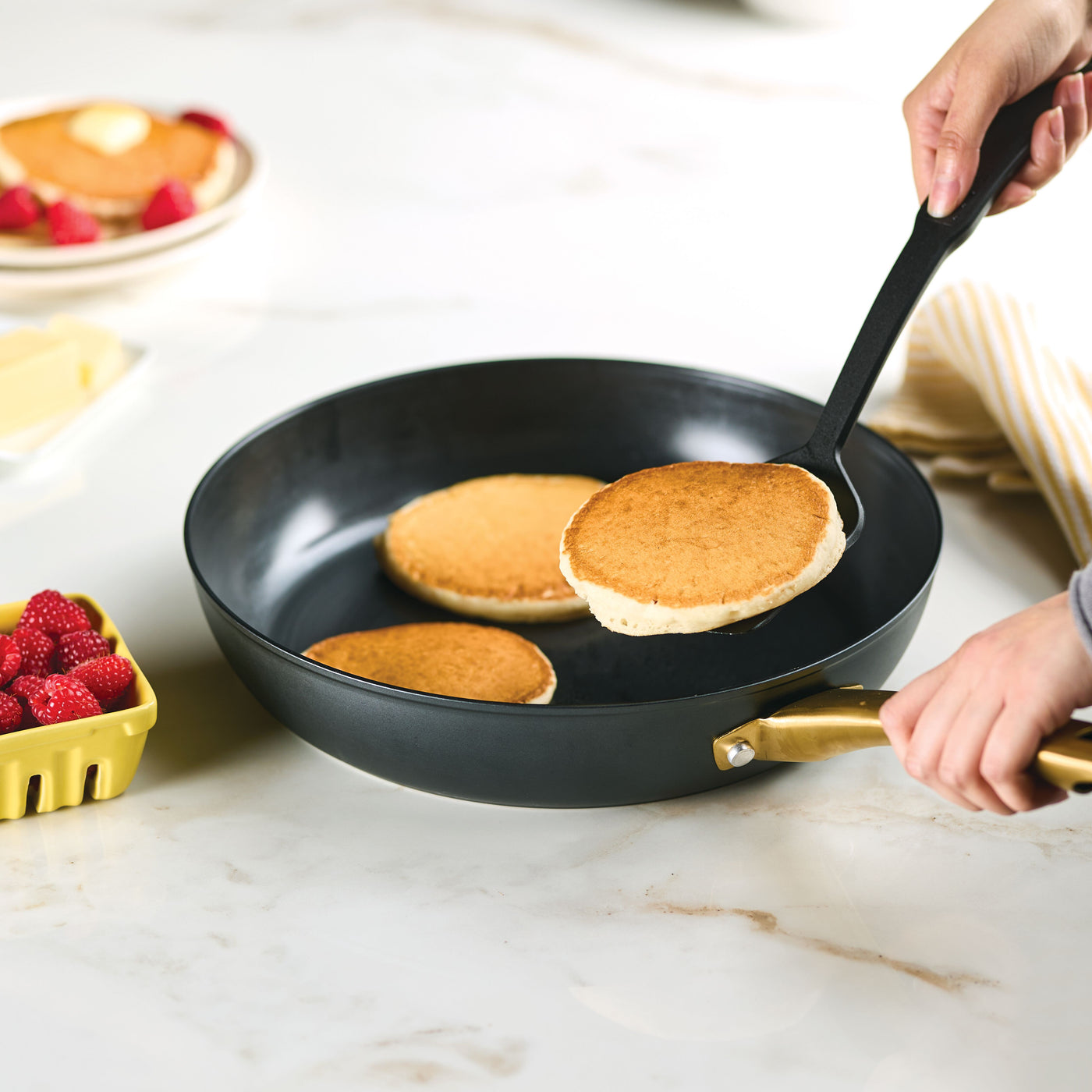 Using the Forged Induction Ceramic 12.25-Inch Frying Pan and a spatula, cook pancakes with raspberries nearby. The induction-ready ceramic nonstick surface allows for effortless flipping, while the forged aluminum with steel bases ensures excellent heat distribution for perfectly golden pancakes every time.