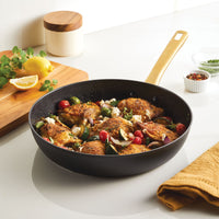 The Forged Induction Ceramic 12.25-Inch Frying Pan, featuring roasted chicken, vegetables, and herbs on a kitchen countertop, is induction-ready and offers even cooking along with easy cleanup.