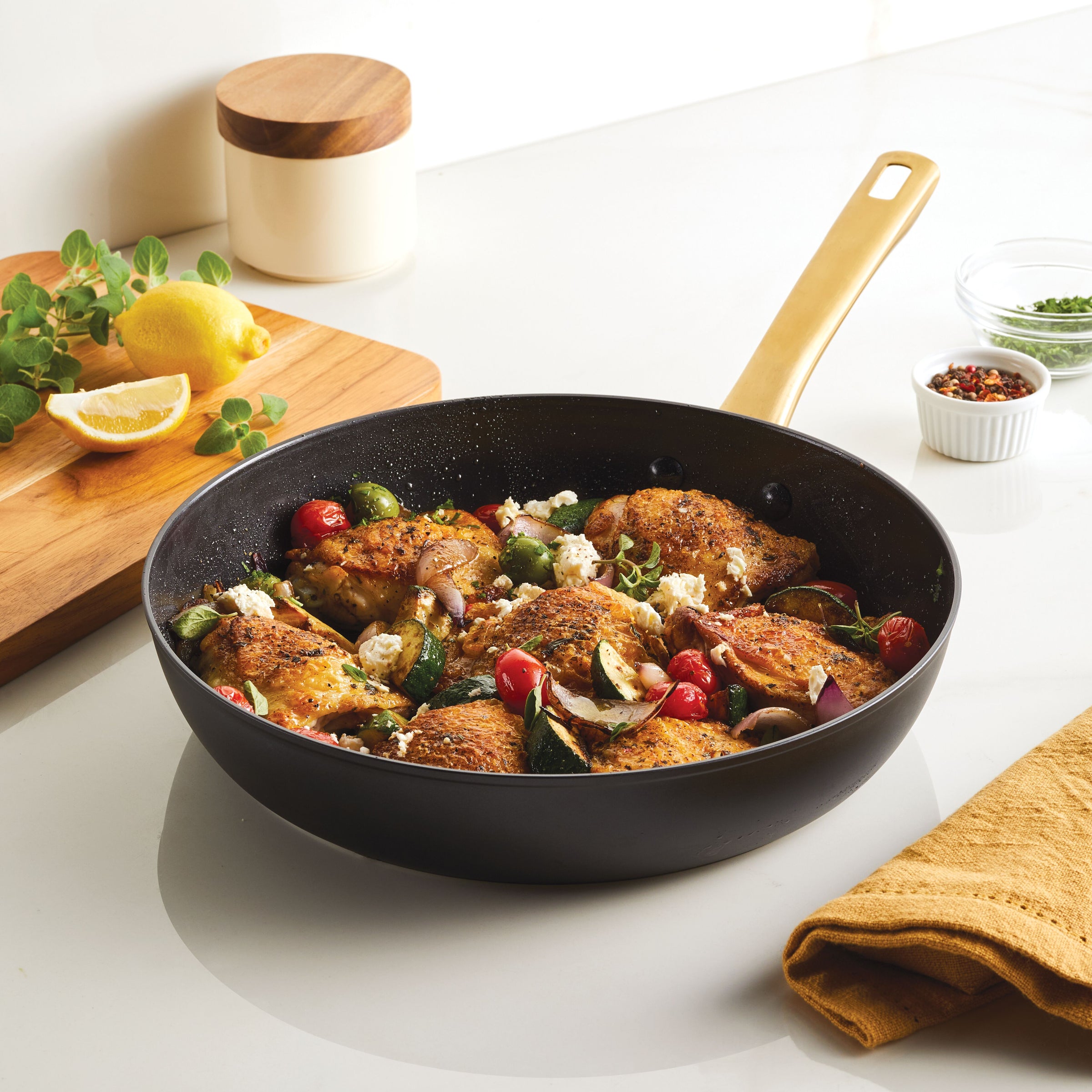 The Forged Induction Ceramic 12.25-Inch Frying Pan, featuring roasted chicken, vegetables, and herbs on a kitchen countertop, is induction-ready and offers even cooking along with easy cleanup.