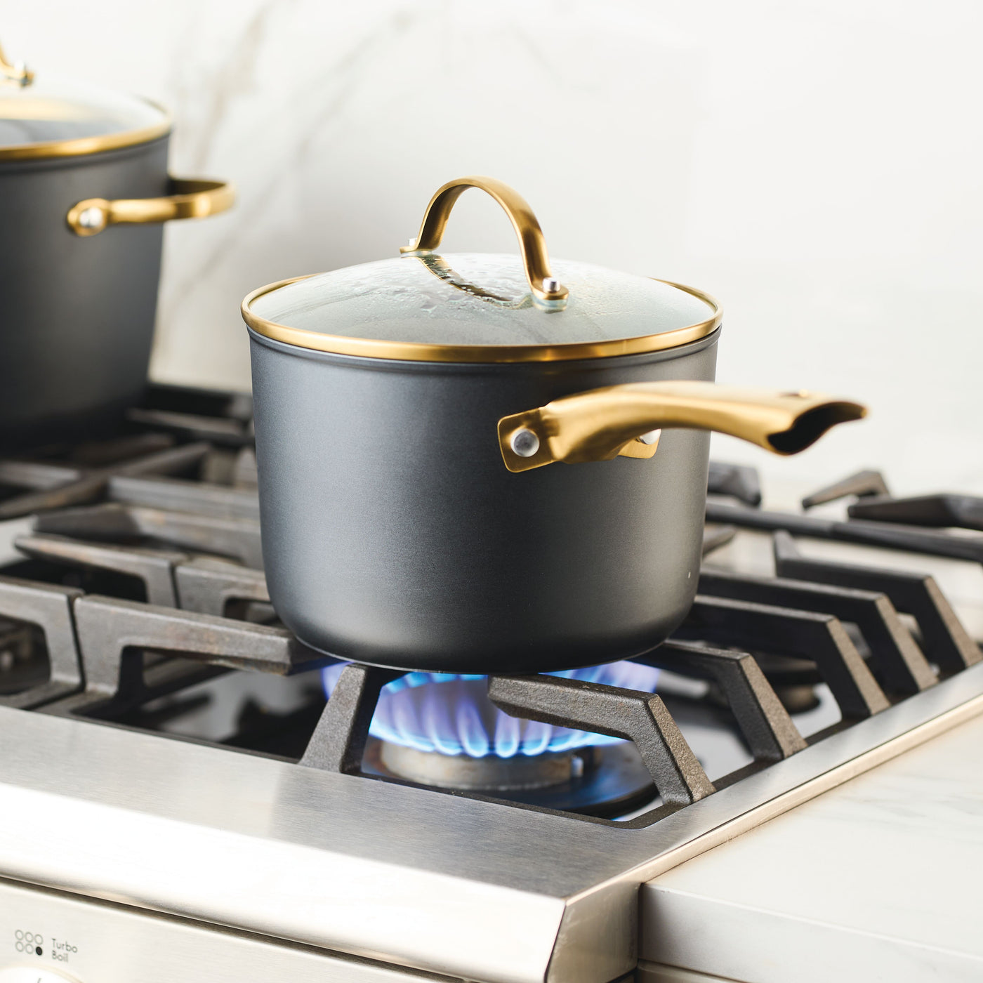 A Forged Induction Ceramic 3-Quart Sauce Pan with a black body and a golden handle rests on a lit gas stove burner.