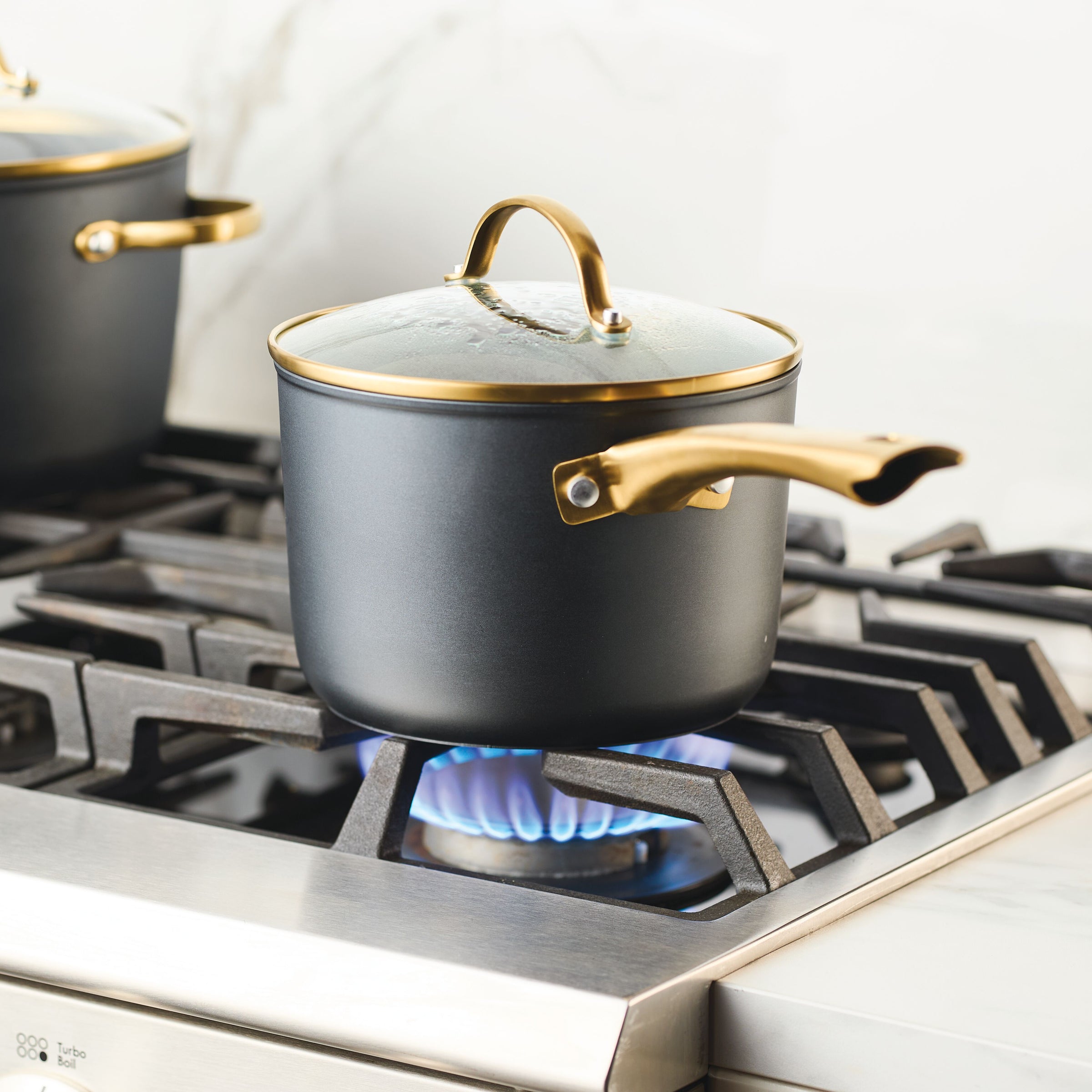 A Forged Induction Ceramic 3-Quart Sauce Pan with a black body and a golden handle rests on a lit gas stove burner.