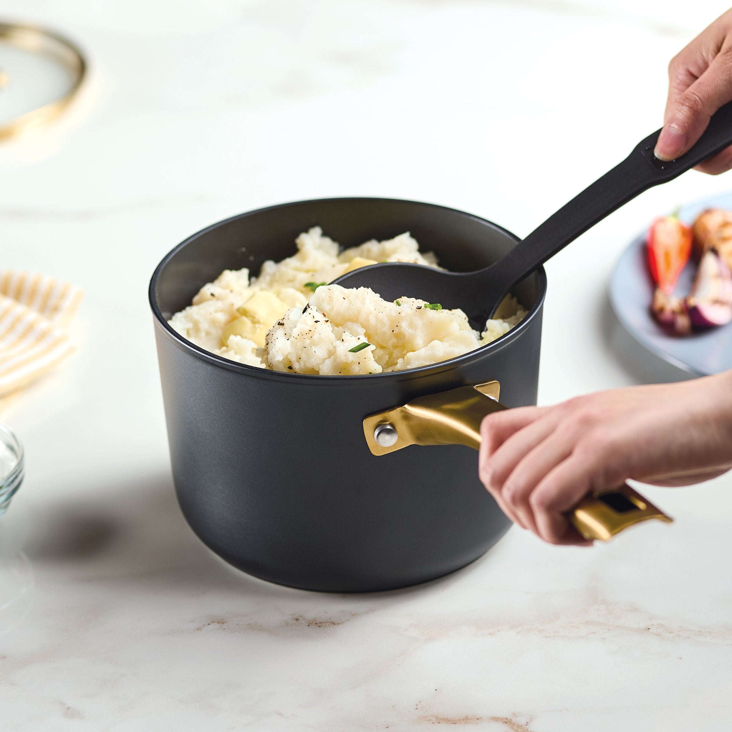 Hands serving mashed potatoes from the Forged Induction Ceramic 3-Quart Sauce Pan With Lid, crafted from forged aluminum with golden handles and featuring induction-ready ceramic nonstick surfaces.