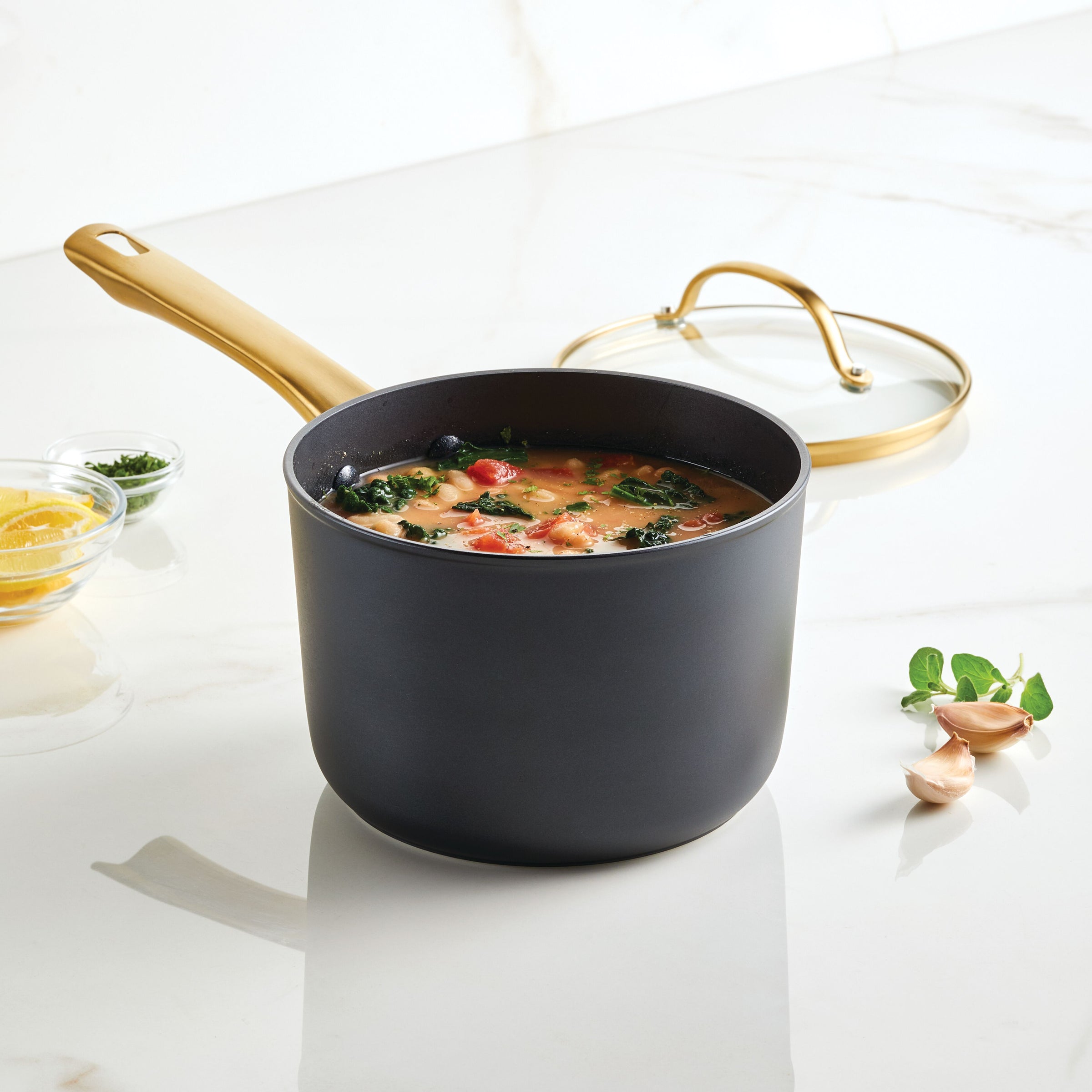 The Forged Induction Ceramic 3-Quart Sauce Pan with Lid, filled with soup and accompanied by garlic and herbs, rests on a marble countertop. It is crafted from forged aluminum and features an induction-ready ceramic nonstick surface.