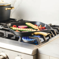 Forged induction ceramic 11-inch square grill pan with vegetables on a gas stove burner.