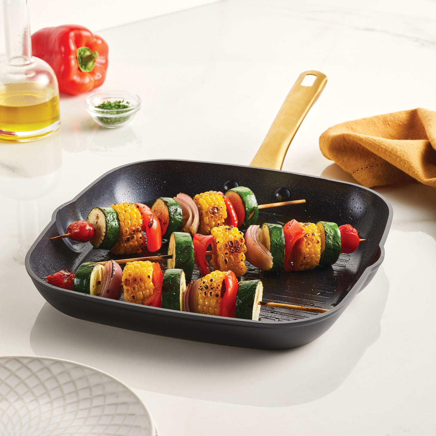 Vegetable skewers sizzle enticingly in the Forged Induction Ceramic 11-Inch Square Grill Pan on the white countertop, ensuring even cooking and effortless cleanup.