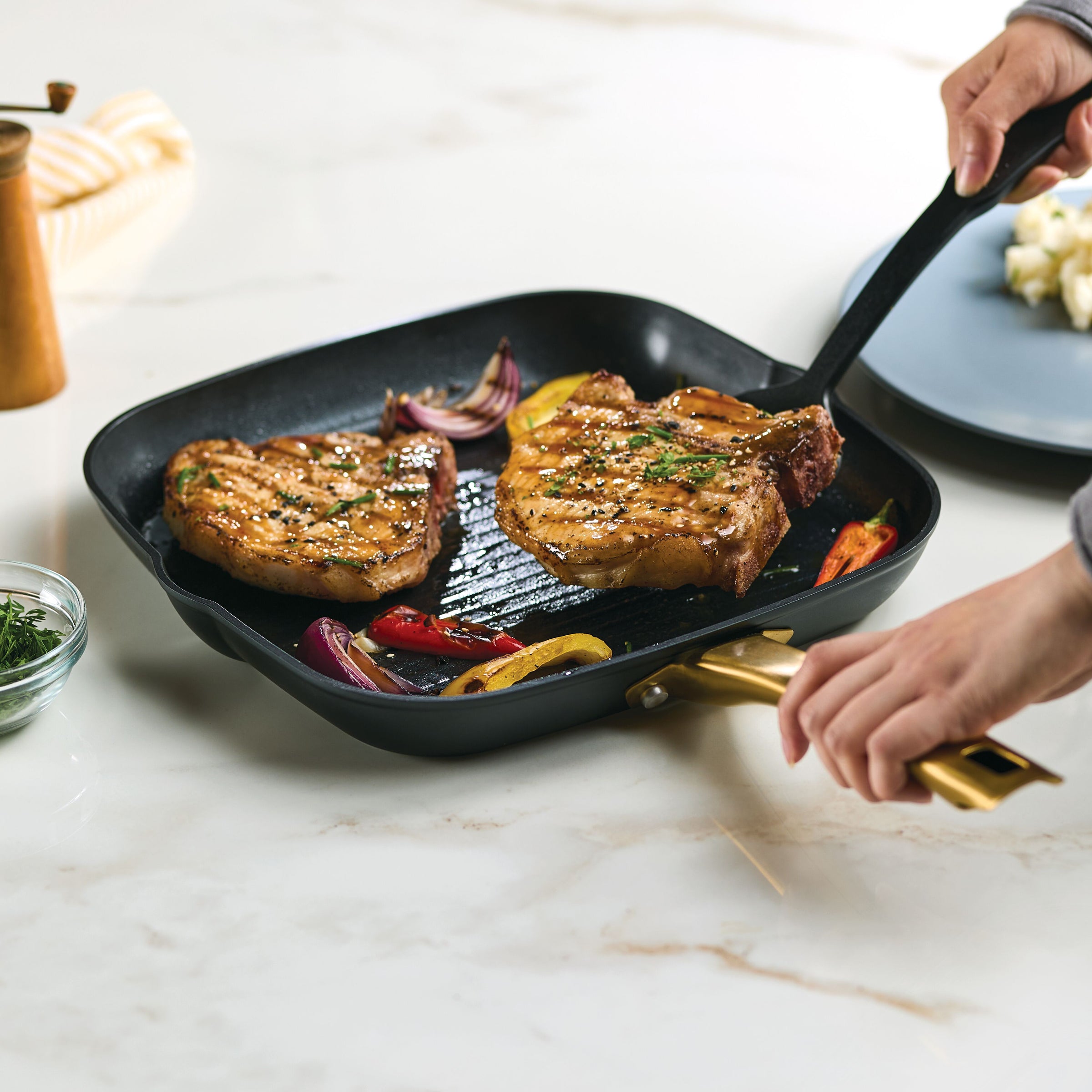 Two pork chops sizzle in a pan with vegetables, cooked to perfection on the Forged Induction Ceramic 11-Inch Square Grill Pan's induction-ready ceramic nonstick surface. The forged aluminum base ensures even heat distribution for a delicious result.