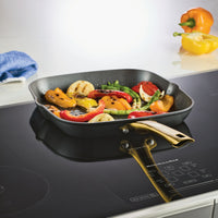 Grilled vegetables in a pan on the Forged Induction Ceramic 11-Inch Square Grill Pan's induction-ready nonstick surface, with bell peppers nearby, sit atop the sleek efficiency of steel bases.