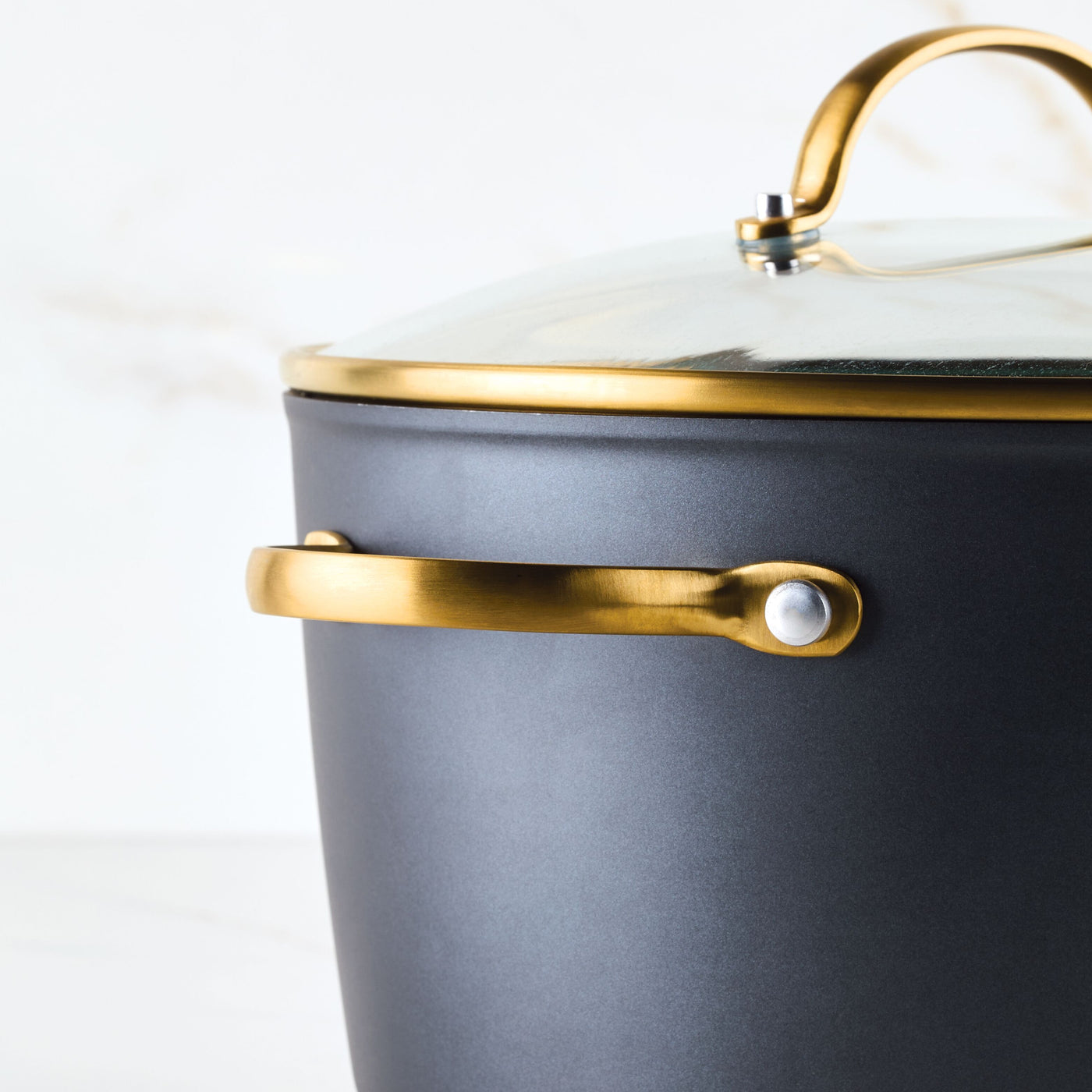 A close-up of the Forged Induction Ceramic 6-Quart Stockpot with a glass lid, featuring gold handles and crafted from durable forged aluminum.
