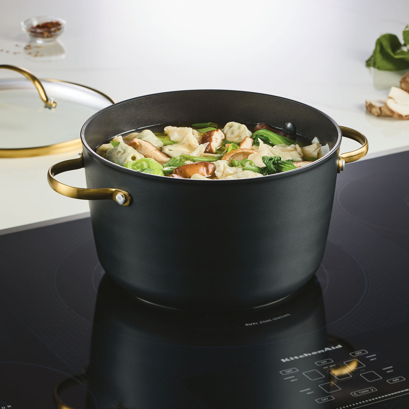 A Forged Induction Ceramic 6-Quart Stockpot With Lid rests on the stovetop, brimming with hearty dumpling soup and fresh vegetables. This induction-ready cookware, crafted from forged aluminum, boasts a ceramic nonstick surface for hassle-free cooking and cleanup.