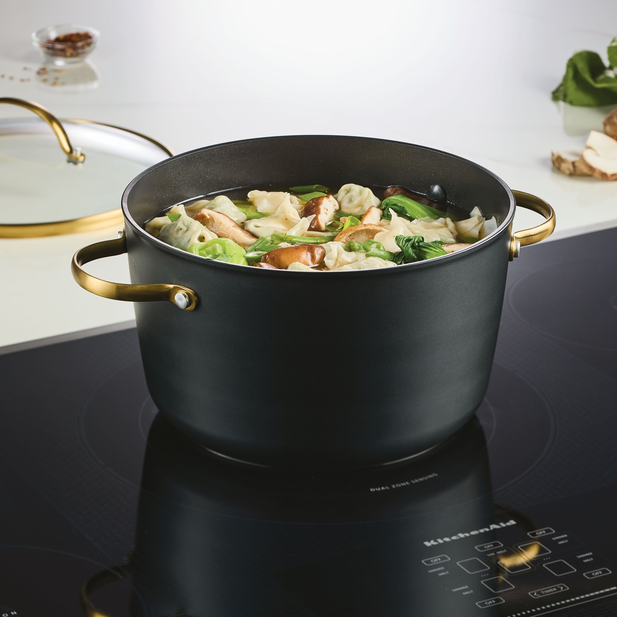 A Forged Induction Ceramic 6-Quart Stockpot With Lid rests on the stovetop, brimming with hearty dumpling soup and fresh vegetables. This induction-ready cookware, crafted from forged aluminum, boasts a ceramic nonstick surface for hassle-free cooking and cleanup.