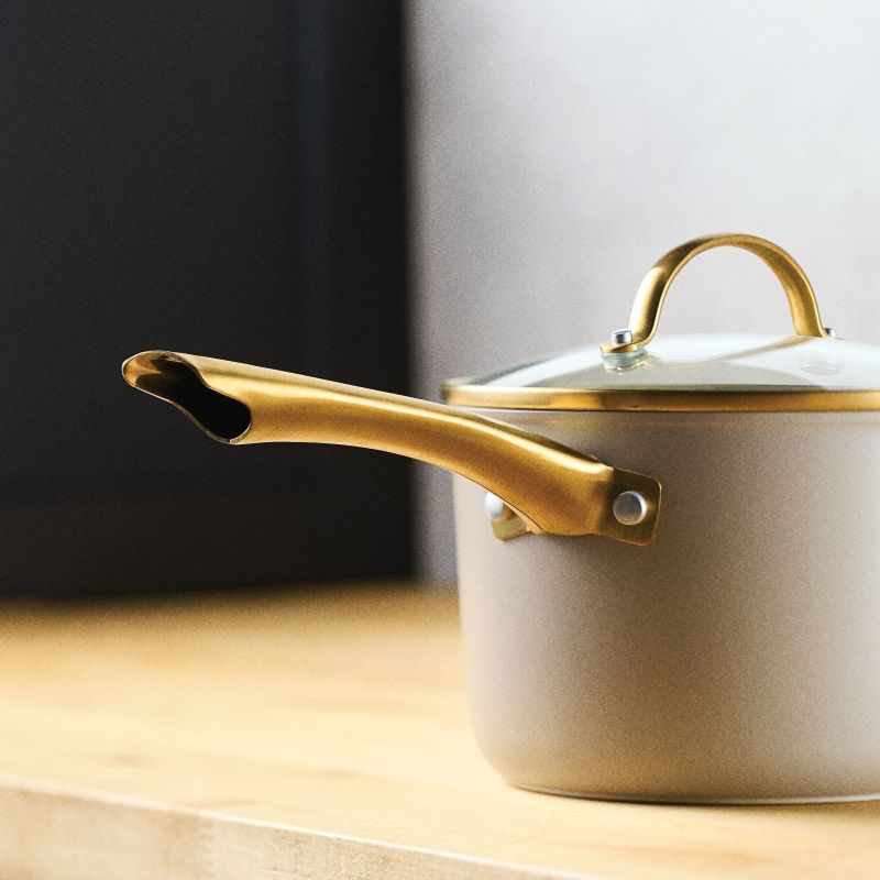 A close-up, side-profile view of the taupe Forged Induction Saucepan  resting on a light wooden countertop. This shot highlights the signature gold-tone handle and the matching gold-tone knob on the glass lid, emphasizing the double-rivet attachment and the smooth matte finish of the taupe exterior.