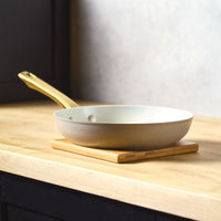 A side-profile view of the Forged Induction Frying Pan in a taupe finish, resting on a wooden trivet. The pan features a cream-colored nonstick interior and a signature gold-tone handle.