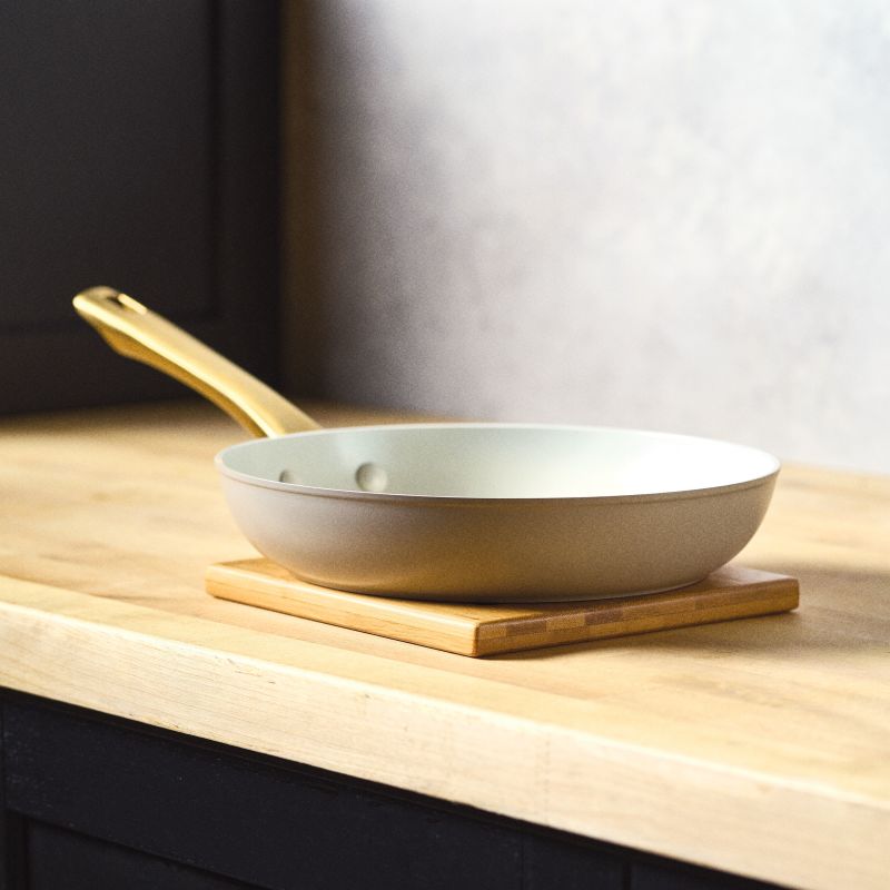 A side-profile view of the Forged Induction Frying Pan in a taupe finish, resting on a wooden trivet. The pan features a cream-colored nonstick interior and a signature gold-tone handle.