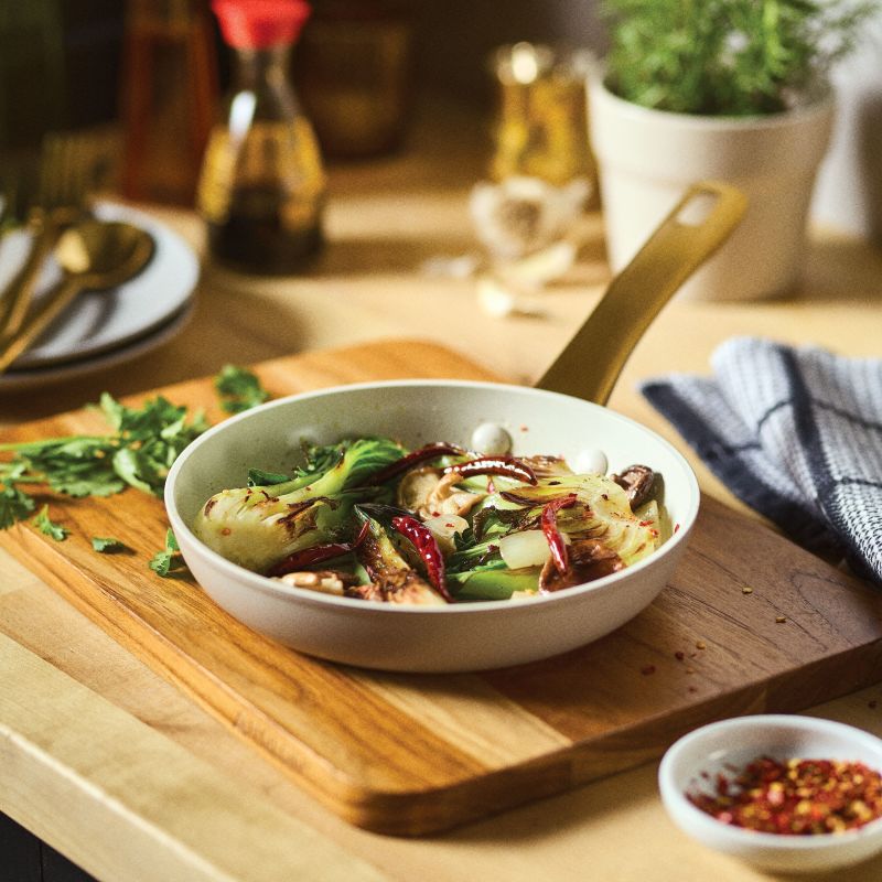 The taupe Forged Induction Frying Pan is shown in a lifestyle setting, filled with a vibrant stir-fry of bok choy, red chilies, and garlic. The pan rests on a wooden cutting board alongside a small bowl of red pepper flakes and a striped kitchen towel, with a blurred kitchen background featuring soy sauce and fresh herbs.