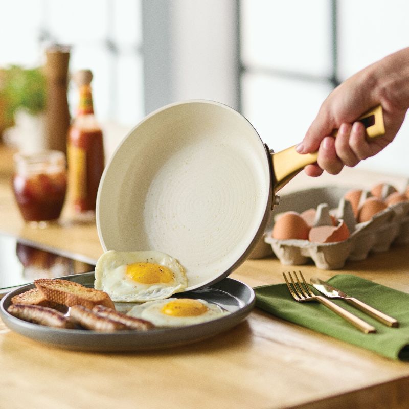 A hand holding the taupe Forged Induction Frying Pan is shown sliding a perfectly fried egg onto a plate. The breakfast spread includes a second fried egg, sausages, and toasted bread on a dark plate, with a carton of eggs and gold-tone flatware resting on a wooden table in the background.