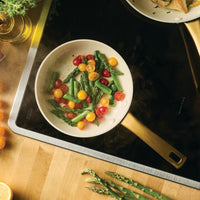 A top-down view of the taupe Forged Induction 8.25-Inch Frying Pan placed on an induction cooktop. The pan is filled with sautéed cherry tomatoes and green asparagus, with sliced lemons and fresh herbs on a wooden cutting board nearby.