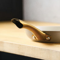 A close-up of the taupe Forged Induction 8.25-Inch Frying Pan focusing on the gold-tone handle's attachment point. The shot highlights the two large, polished rivets securing the handle to the side of the pan and the smooth, matte finish of the taupe exterior.