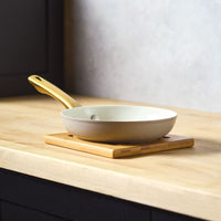 A side-profile view of the Forged Induction 8.25-Inch Frying Pan in a taupe finish, resting on a wooden trivet. The pan features a cream-colored nonstick interior and a signature gold-tone handle.