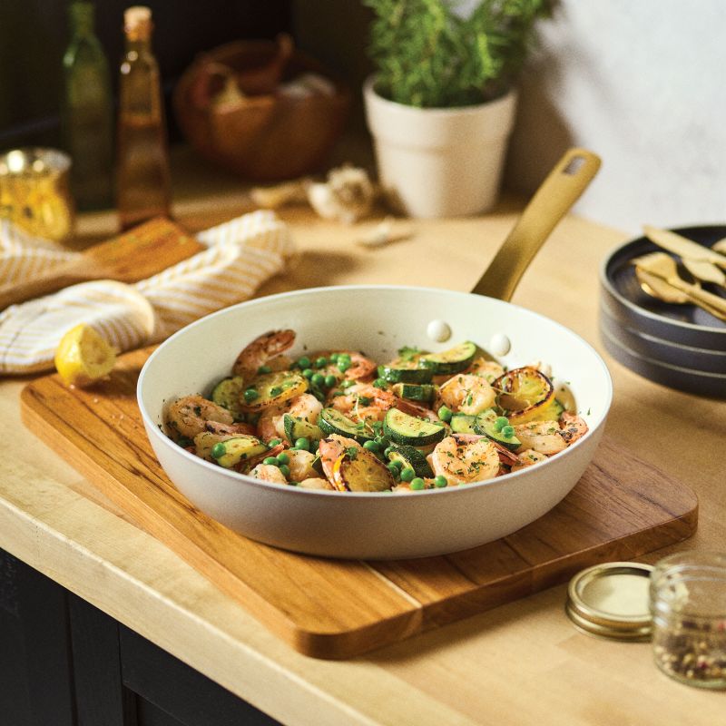 The taupe 12.25-Inch Forged Induction Frying Pan is shown in a lifestyle setting, resting on a wooden cutting board. The pan is filled with a Mediterranean-style sauté of shrimp, sliced zucchini, peas, and charred lemon rounds. In the background, a light-colored kitchen counter holds a potted herb plant, garlic cloves, and several glass bottles.